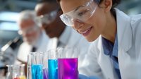 Young female scientist in protective eyewear examining colorful liquid samples in laboratory glassware while working with diverse research team in modern facility.