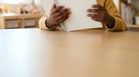Woman with dark skin tone wearing a yellow cardigan and beaded bracelet prays at wooden table in home interior with blurred background.