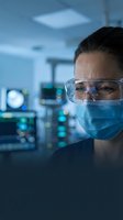 Female scientist wearing protective mask and safety glasses in modern laboratory with medical equipment and blue lighting for research work.