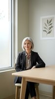 Mature woman with white hair, Caucasian ethnicity, wearing black blazer and beige blouse, seated at wooden desk in bright modern office with large window and botanical wall art.