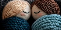 Knitted doll faces touching foreheads tenderly, wrapped in cozy blue yarn. Close-up macro shot showing detailed textile texture and embroidered sleeping expressions.