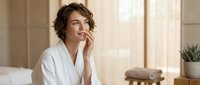Young woman with short wavy brown hair and fair complexion wearing white robe, sitting on bed in bright bedroom, hand near face in thoughtful pose, morning skincare routine.