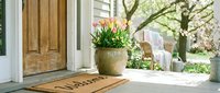 Cozy front porch with a wooden door, welcome mat, and colorful tulips in a stone pot, surrounded by blooming spring trees.