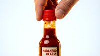 Unidentified male hand holding red-capped hot sauce bottle with habanero heat label against white background for condiment marketing.