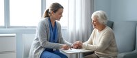 Young female healthcare professional in scrubs consulting with elderly Caucasian woman patient in bright medical office setting for compassionate care.
