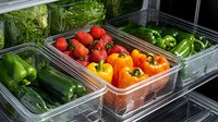 Fresh organic produce in refrigerator drawers with strawberries, bell peppers, and cucumbers arranged in clear storage containers for healthy meal preparation.