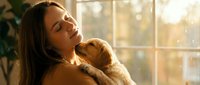 Young woman cuddling adorable golden retriever puppy in warm sunlit room by window. Tender moment of pet bonding and companionship at home.
