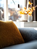 Cozy knitted mustard yellow cushion on gray sofa with soft bokeh fairy lights in background creating warm autumn atmosphere for home interior styling.