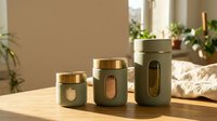 Modern sage green kitchen canisters with gold lids and viewing windows on wooden counter in sunlit home interior for food storage organization.