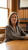 Mature Caucasian female attorney in gray blazer seated at wooden desk in law office with legal books and diploma visible in background.