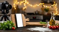 Food blogger setup with smartphone on wooden stand, camera on tripod, fresh ingredients and warm bokeh lights for cooking video content creation.