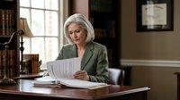 Mature Caucasian businesswoman in green blazer reviewing documents at wooden desk in traditional office setting with natural window light.