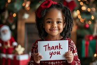 Young African American girl with curly hair and red bow holding thank you sign, wearing festive dress against Christmas tree background with bokeh lights and decorations.