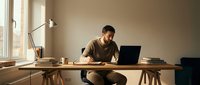 Young man with dark hair and beard, wearing casual brown shirt, focused on work at wooden desk with laptop, coffee cup, and desk lamp in minimalist home office space.