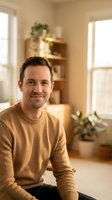 Smiling middle-aged Caucasian man in beige sweater sitting in bright modern living room with plants and natural light for lifestyle content.
