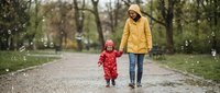 Mother and toddler daughter walking hand in hand on park path during spring rain, wearing bright yellow and red raincoats for outdoor family activities.