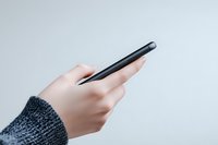 Female hand in gray knitted sweater holding black smartphone against light background, minimalist composition showing modern technology use in daily life.