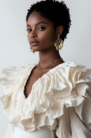 Young African American woman wearing cream ruffled designer blouse with dramatic pleated collar, gold statement earrings and necklace against light background. Natural curly hair.