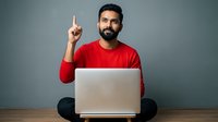 Young bearded Middle Eastern man in red shirt pointing up with finger while working on laptop computer, having creative idea or eureka moment.