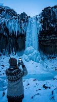 Rear view of person wearing knit sweater and beanie photographing frozen waterfall cascading between basalt columns in winter landscape.