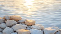 Smooth river rocks stacked on water edge at sunset, golden sunlight reflecting on gentle ripples creates serene natural meditation scene for wellness background.