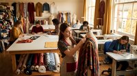 Young female fashion designer examining burgundy fabric in bright textile studio workspace with colorful materials and creative team working in background.