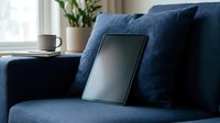 Tablet with blank screen leaning on navy blue sofa cushions in cozy modern living room, coffee cup and plant in background.