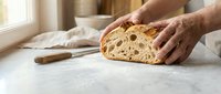 Hands slicing fresh artisan sourdough bread on marble counter in bright kitchen. Homemade baking and cooking concept for food preparation and culinary arts.