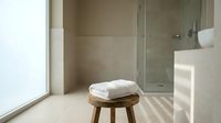 Modern minimalist bathroom interior with wooden stool, white towels, glass shower, natural light creating shadows on beige tiles for spa design.