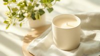 Aromatic soy candle in white ceramic holder with natural shadows and green plant decoration on light linen fabric, creating cozy atmosphere.