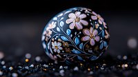 Decorative black sphere with delicate pink floral pattern and blue leaves painted on surface, placed on dark background with glittering particles.