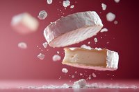 Floating pieces of soft cheese with white rind and creamy interior against pink background with scattered crumbs in dynamic composition for food industry.