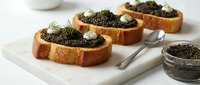 Toasted bread slices topped with creamy spread, caviar, and fresh dill garnish arranged on white plate with serving spoon and caviar jar, suitable for gourmet appetizers and fine dining presentations.