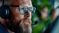 Caucasian male customer service representative with beard and glasses wearing headset in office environment. Professional call center agent focused on work.