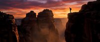 Silhouetted figure standing on rocky cliff edge at dramatic sunset with golden and orange sky over misty canyon landscape, conveying adventure and exploration themes.