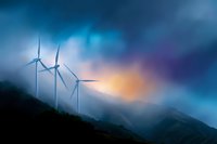 Wind turbines silhouetted against dramatic sky at sunset, with mountains in background and moody blue storm clouds creating atmospheric landscape.