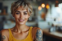 Young blonde woman with short pixie haircut and tattoos smiling warmly in casual yellow tank top at modern cafe or restaurant setting.