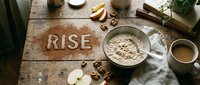 Oatmeal breakfast flatlay on rustic wooden table with word RISE written in cinnamon powder, walnuts, apple slices, and coffee cup.