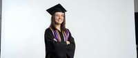 Young Caucasian female graduate in black cap and gown with colorful honor cords smiling confidently with crossed arms on white background.