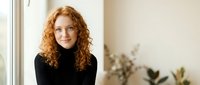 Professional redhead woman with curly hair in black turtleneck smiling confidently by window in modern office interior for business and career concepts.