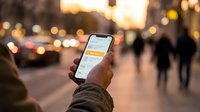 Man holding smartphone with buy now button on screen, standing on busy city street at dusk with blurred pedestrians and traffic lights.