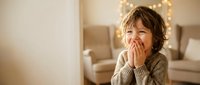 Adorable toddler boy with curly hair covering mouth with hands in cozy living room with warm lighting. Child expressing surprise, shyness or playfulness at home.