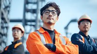 Hispanic male construction worker in orange safety vest and glasses with confident pose, team members in background at industrial site. Professional workplace portrait.