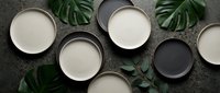 Empty ceramic bowls and plates in white and black arranged on gray concrete surface with green tropical leaves, flat lay composition for tableware and dinnerware design projects.