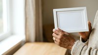 Senior person with brown skin holding empty white picture frame indoors by window, displaying blank space for artwork or family photos.
