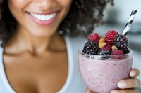 Young African American woman holding pink berry smoothie bowl topped with fresh blackberries, raspberries and nuts with striped straw in modern kitchen.