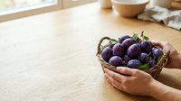 Fresh purple eggplants in wicker basket held by hands on wooden table with natural light, suitable for food, cooking, and agricultural content.