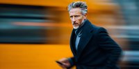 Mature businessman in dark coat walking quickly against blurred yellow urban background, holding smartphone, conveying urgency and city lifestyle.