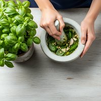 Hands preparing fresh basil pesto in stone mortar with pestle, traditional Italian cooking method for homemade sauce preparation on wooden kitchen counter.