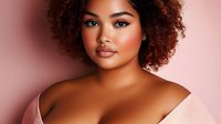 Young African American woman with natural curly hair and glowing skin against pink background, wearing off-shoulder sweater, expressing confidence and beauty.